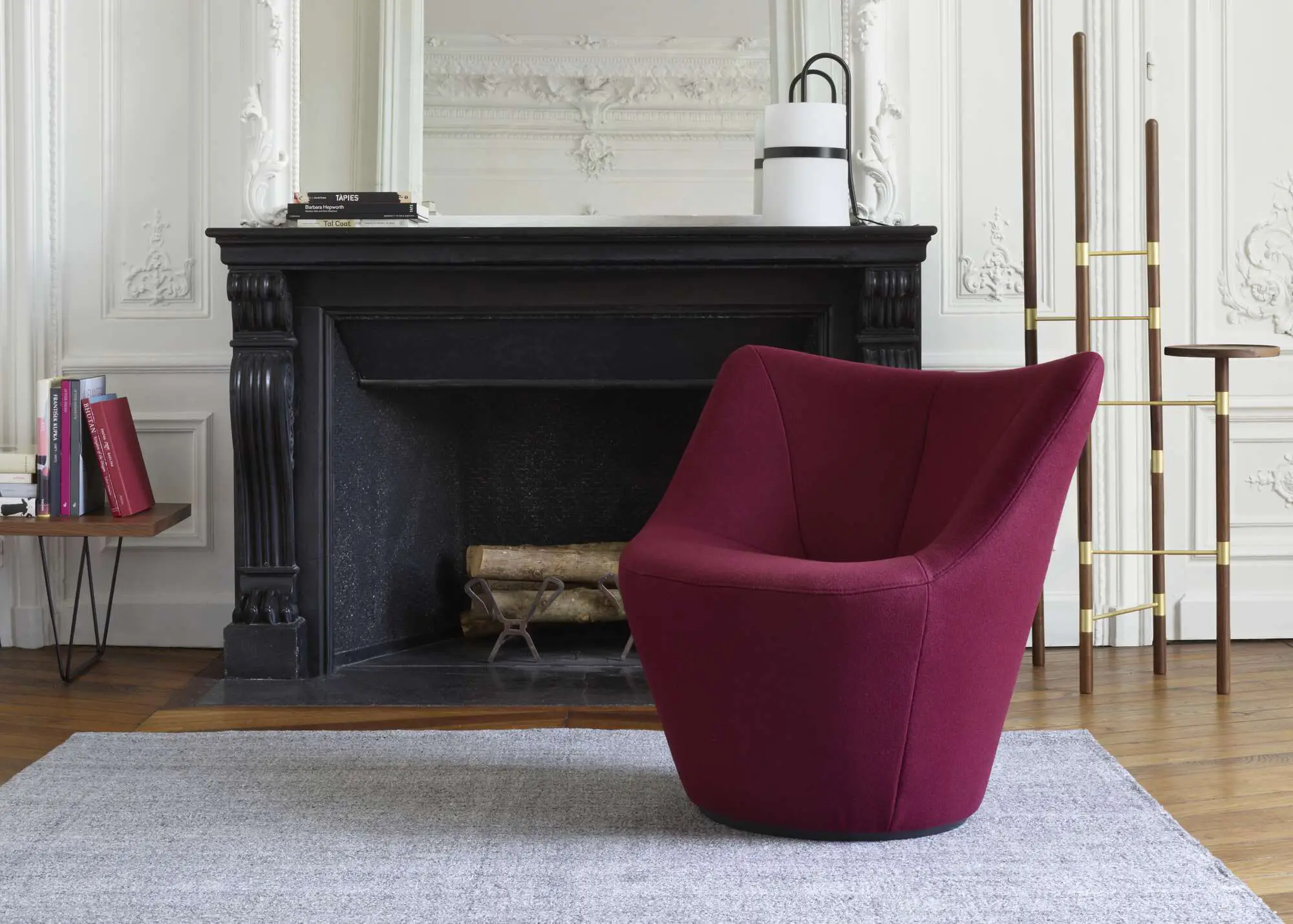 Modern maroon armchair on a gray rug in front of a black fireplace with wood logs, flanked by a side table with books and a tall wooden coat rack.