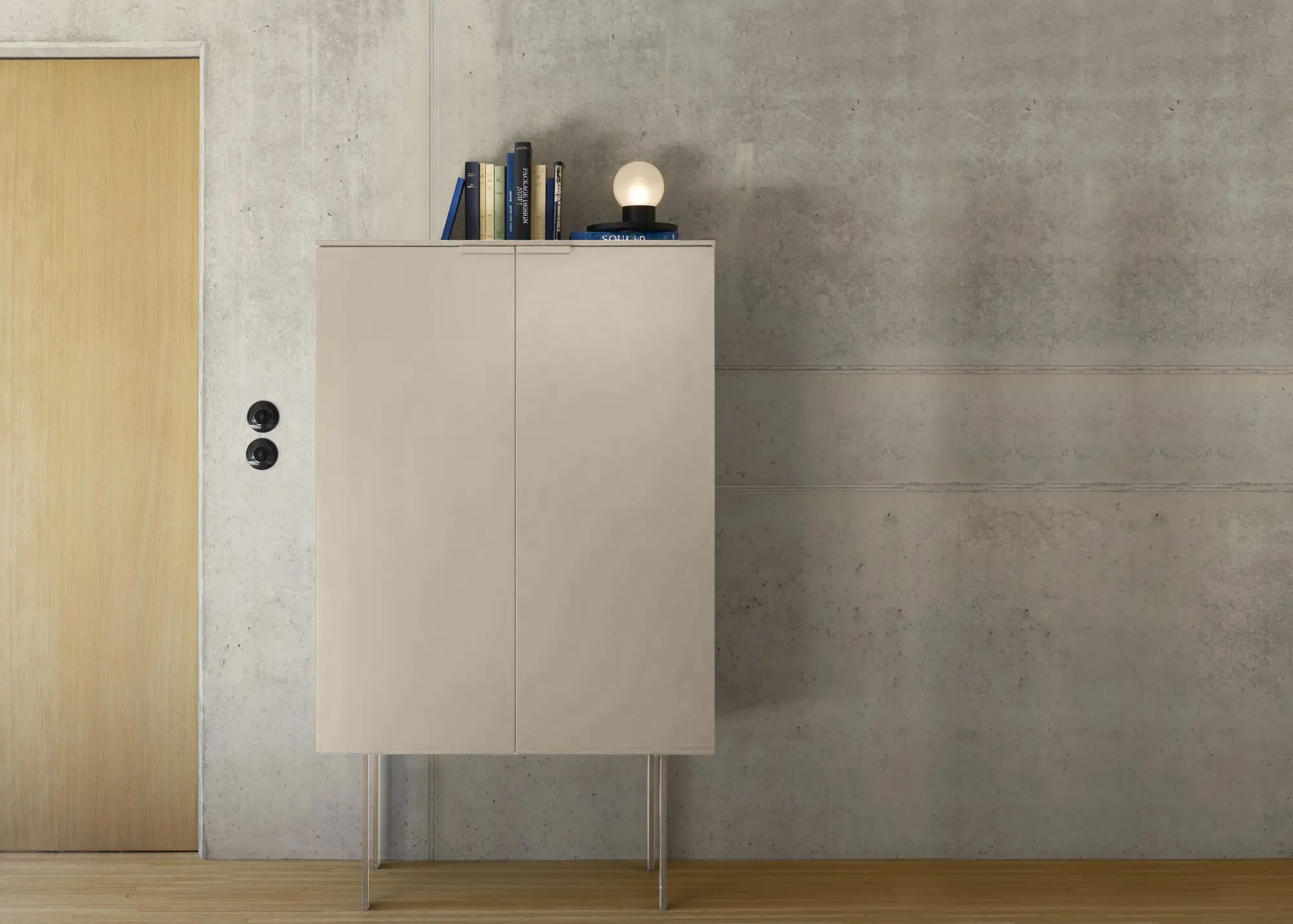 Minimalist beige cabinet with slim legs against a concrete wall, topped with books and a small round lamp.