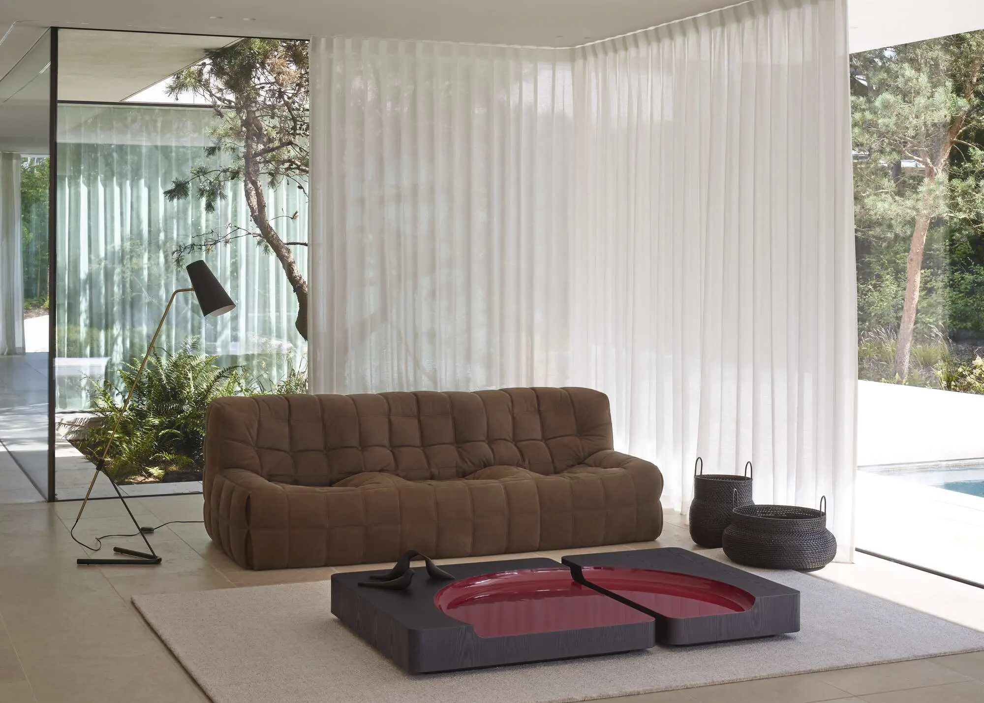 Modern living room with a brown tufted sofa, a black floor lamp, and a unique low coffee table with red circular inset, near large glass windows with sheer white curtains.