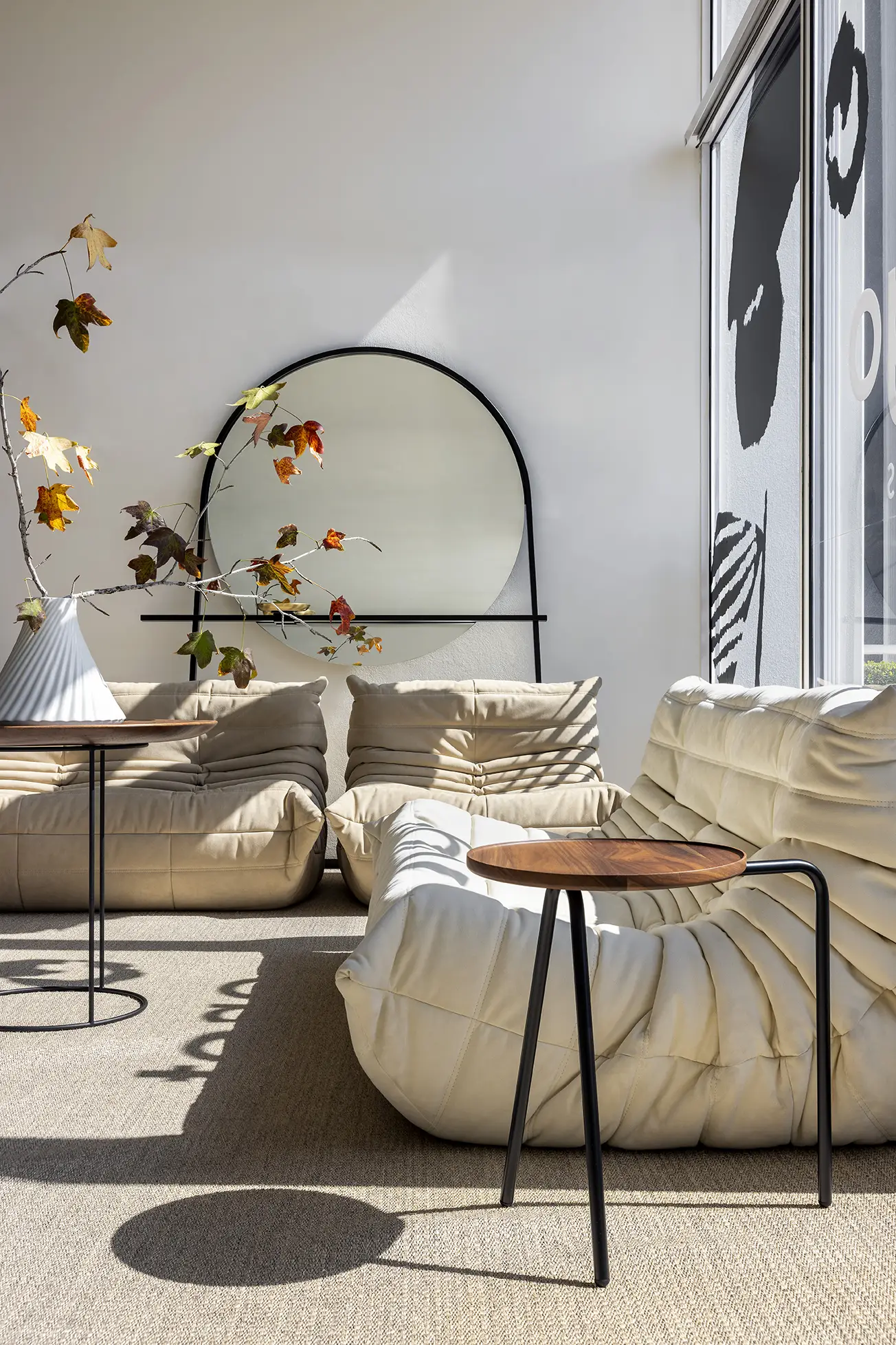 Modern beige lounge chairs with a round wooden side table and a white vase with autumn leaves in a sunlit room with a round black-framed mirror on the wall.