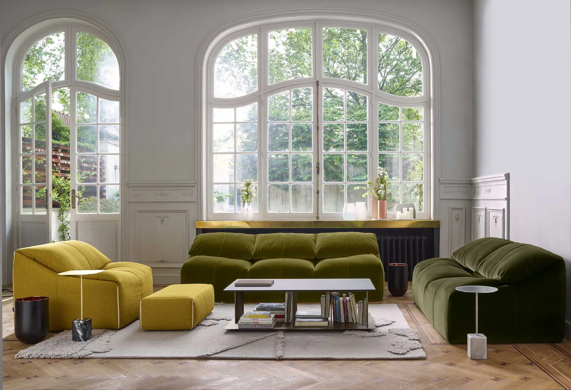 Modern living room with large arched windows, green and yellow plush sofas, a low coffee table with books, and decorative plants.