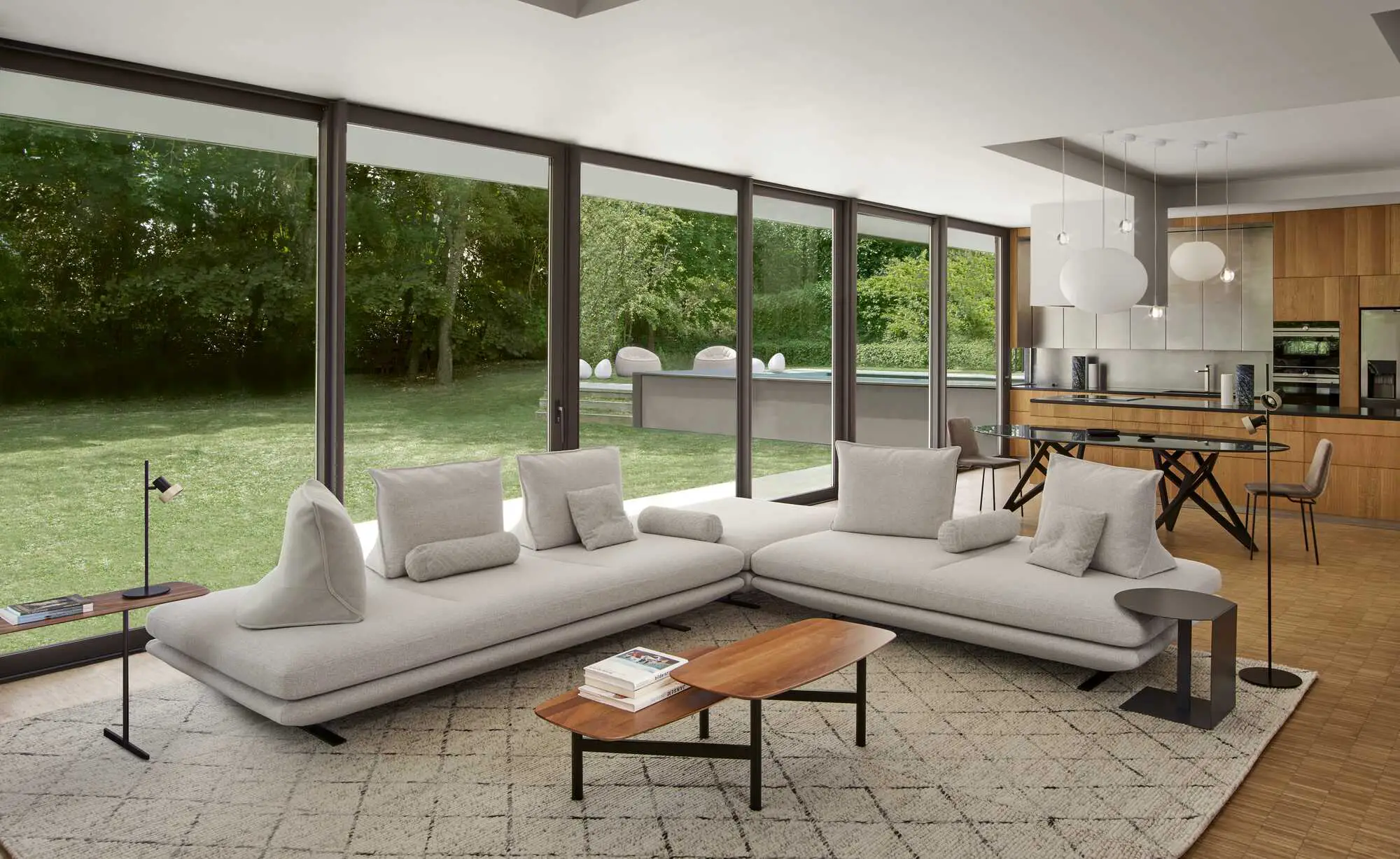 Modern living room with large beige sectional sofa, wooden coffee table, floor lamps, and floor-to-ceiling windows overlooking a green backyard with a pool.