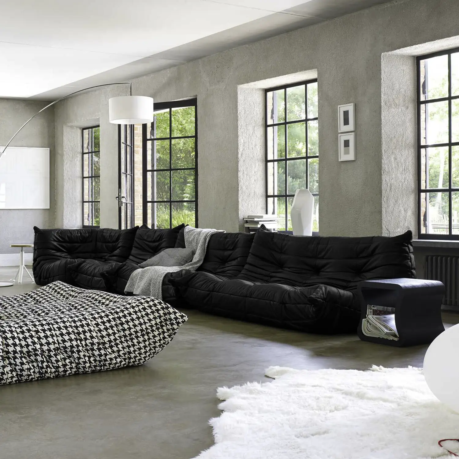 Modern living room with large black sectional sofa, patterned ottoman, arched floor lamp, and large windows with views of green trees.